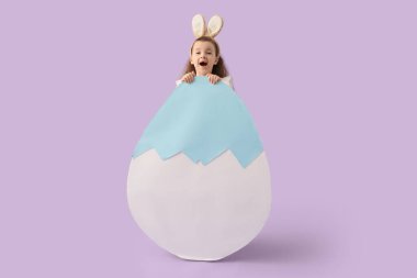Şaşkın küçük kız tavşan kulaklı Büyük kağıt Paskalya yumurtası ile leylak arka planda