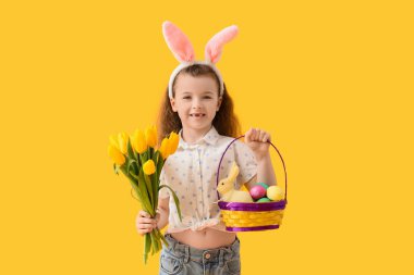 Paskalya yumurtası sepeti ve sarı arka planda laleleri olan tavşan kulaklı şirin bir kız.