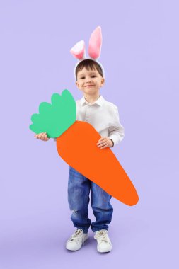 Leylak arka planında büyük kağıt havuç olan tavşan kulaklı şirin bir çocuk. Paskalya kutlaması
