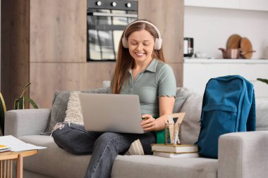 Kulaklıklı, dizüstü bilgisayarlı genç kız evdeki kanepede çalışıyor.