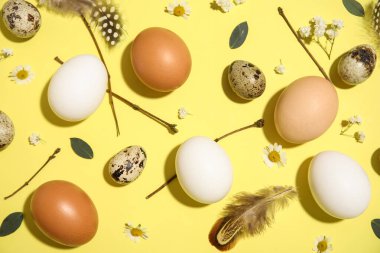 Sarı arka planda Paskalya yumurtaları, çiçekler, tüyler ve dallarla kompozisyon