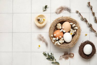 Paskalya yumurtaları, çiçekler, tüyler ve beyaz fayanslı arka planda söğüt dalları olan yuva.