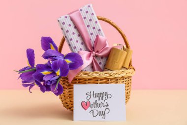 Üzerinde süsen çiçekleri olan sepet, Anneler Günü için hediye kutusu ve kozmetik ürünleri pembe arka planda bej masada.