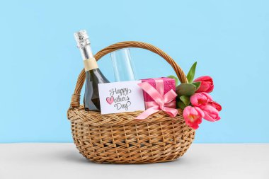 Laleli sepet, şampanya şişesi ve mavi arka planda beyaz masanın üzerinde hediye kutusu.