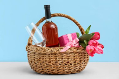 Laleli sepet, şarap şişesi ve Anneler Günü için hediye paketi mavi arka planda beyaz masada.