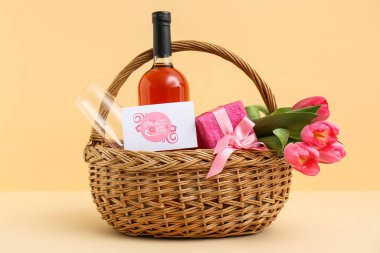 İçinde laleler, şarap şişesi ve bej arka planda anneler günü için hediye paketi olan sepet.