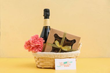 Sarı arka planda Anneler Günü için karanfilli sepet, şampanya şişesi ve çikolata şekeri.