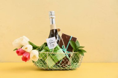 Sarı zemin üzerinde şampanya şişesi, lale ve çikolata olan sepet.
