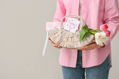 Beyaz arkaplanda anneler günü için içinde laleler, kartpostal ve hediye kutusu olan sepeti tutan genç bir kadın.