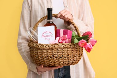 Sarı arka planda Anneler Günü için elinde şarap şişesi, lale ve hediye kutusu olan genç bir kadın var.