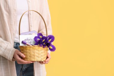 Sarı arkaplanda anneler günü için çiçek, kartpostal ve kozmetik taşıyan sepet taşıyan genç bir kadın.
