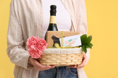 Sarı arka planda Anneler Günü için karanfilli, şampanya şişeli ve çikolatalı sepet taşıyan genç bir kadın.