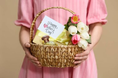 Bej arka planda Anneler Günü için hediye sepeti, kozmetik ve lale taşıyan genç bir kadın.