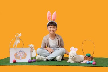 Paskalya sepetleri, tebrik kartları ve sarı arka planda yeşil çimlerde oturan beyaz kediyle tavşan kulaklı şirin bir çocuk.