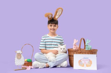 Beyaz kedisi, Paskalya sepeti ve leylak arka planında tebrik kartı olan tavşan kulaklı şirin bir çocuk. Paskalya kutlaması