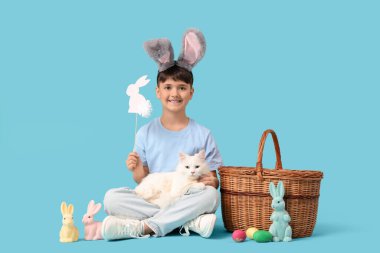 Tavşan kulaklı şirin bir çocuk, oyuncak tavşanlar, paskalya yumurtaları ve mavi arka planda beyaz kedi.