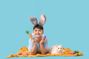 Oyuncak havuç, paskalya yumurtası ve mavi arka planda yatan beyaz kediyle tavşan kulaklı şirin bir çocuk.