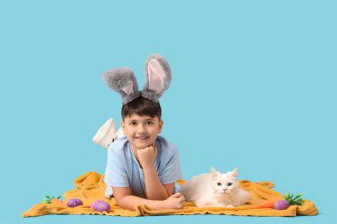 Oyuncak havuç, paskalya yumurtası ve mavi arka planda yatan beyaz kediyle tavşan kulaklı şirin bir çocuk.