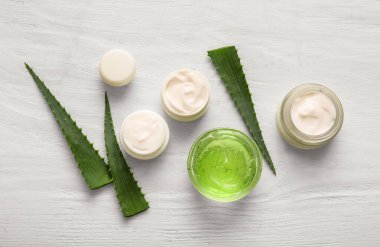 Kozmetik krem kavanozları ve beyaz ahşap arka planda aloe vera yapraklı cam kase.