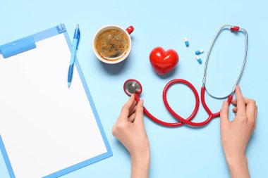Steteskop, kırmızı kalp, bir fincan kahve ve mavi arka planda pano olan kadın elleri. Dünya Sağlık Günü