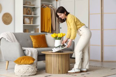 Oturma odasındaki sehpanın üstünde lale çiçekleri ve dergilerle dolu vazosu olan mutlu genç bir kadın.