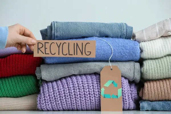 Masadaki giysi yığınlarının yanında RECYCLING tabelası tutan kadın, yakın plan.