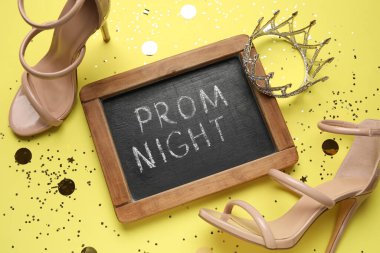 SMS PROM gecesi yazılı tebeşir tahtası, yüksek topuklu ayakkabı, taç ve sarı arka planda konfeti.