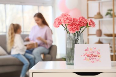 İçinde karanfil ve kartpostal olan vazo, Anneler Günü için masada, kızı oturma odasında olan genç bir kadına karşı, yakın plan.