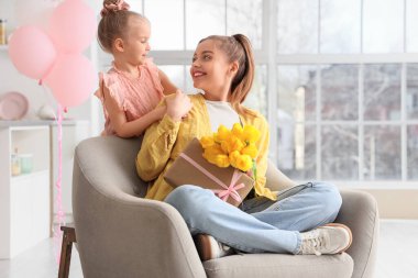 Anneler Günü 'nde sarı laleler ve evdeki koltukta hediye kutusuyla annesini selamlayan tatlı küçük kız.