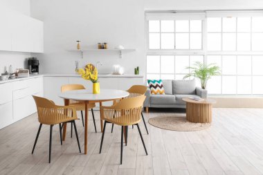 Yemek masası, mimoza çiçekleri, koltuklar, ev bitkisi, tezgah ve kanepesi olan ferah mutfak stüdyosunun içi.