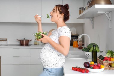 Genç hamile kadın mutfakta taze sebze salatası yiyor.