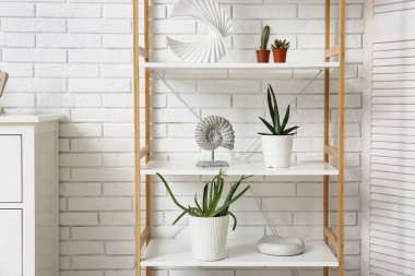 Beyaz tuğla duvarın yanında yeşil bitkiler ve dekor bulunan raf ünitesi.