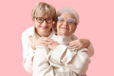 Olgun bir kadın pembe arka planda annesine sarılıyor.