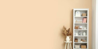 Kitaplarla, vazolarla ve bej duvarın yanındaki dekorasyonlarla dolu raf ünitesi. Tasarım için pankart