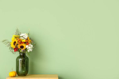 Yeşil duvarın yanında bir buket sonbahar çiçeği ve balkabağı olan raf ünitesi.