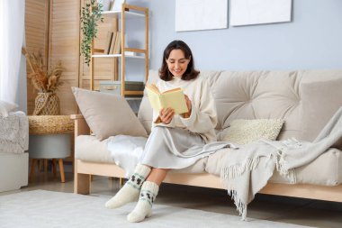 Sıcak kazaklı genç bir kadın evdeki kanepede kitap okuyor.