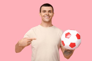 Yakışıklı genç adam pembe arka planda futbol topuna işaret ediyor.