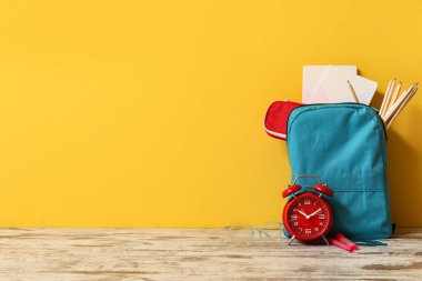 Sarı duvarın yanındaki ahşap masada mavi okul çantası ve alarm saati.