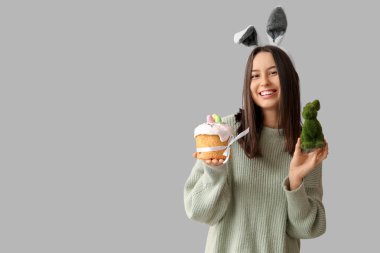 Tavşan kulaklı güzel bir genç kadın, oyuncak tavşan ve gri arka planda Paskalya pastası.