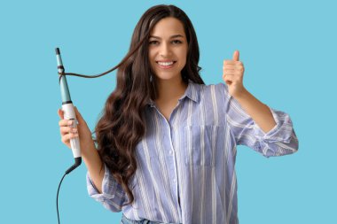 Mavi arka planda baş parmağını gösteren saç maşasıyla güzel bir genç kadın.