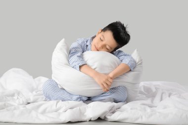 Beyaz arka planda pijamalı, yastıklı ve battaniyeli uykulu küçük Asyalı çocuk oturuyor.