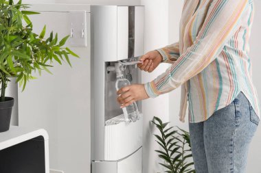 Plastik şişeyi beyaz duvarın yanındaki soğutucudan filtreli suyla dolduran kadın.