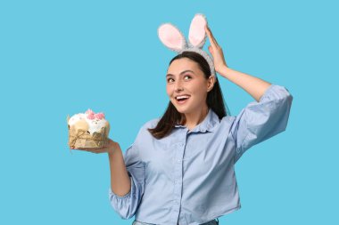 Mavi arka planda Paskalya pastası olan tavşan kulaklı güzel genç bir kadın.