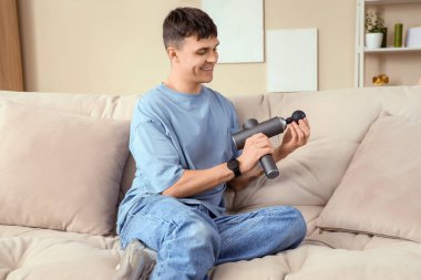 Evde kanepede oturan vurmalı masaj aleti olan genç bir adam.