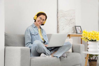 Kulaklıklı genç bir kadın evdeki kanepede dizüstü bilgisayar kullanıyor.