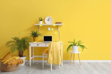 Çalışma odası, dizüstü bilgisayar, sandalye ve saat sarı duvarda.