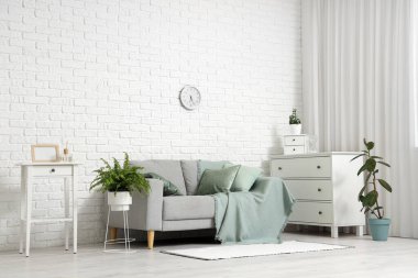 Gri kanepe, ev bitkileri, çekmeceler ve şık oturma odasındaki beyaz tuğlalı duvar üzerindeki saat.