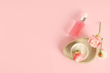 Bir şişe kozmetik yağı, güller, kase ve taş tepsi pembe arka planda.