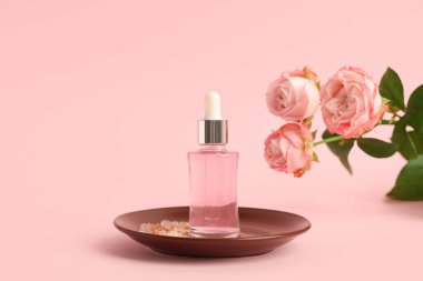 Bir şişe kozmetik yağı, güller ve pembe arka planda deniz tuzu olan bir fincan tabağı.