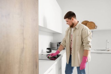 Genç sakallı adam mutfakta elektrikli sobayı süngerle temizliyor.